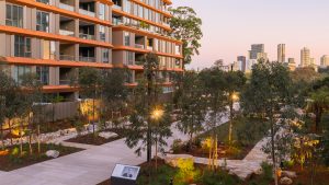 Henderson Park, adjacent to a contemporary apartment building, offers pathways and trees under a clear evening sky. With the city skyline in the background, this modern urban park hosts events like the official opening of NINE by Mirvac.