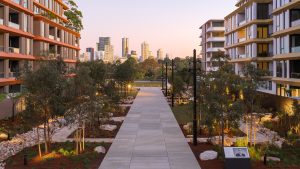 The newly unveiled Henderson Park features a modern urban landscape with a wide paved walkway lined with trees and greenery. On either side are contemporary apartment buildings, such as NINE by Mirvac. In the distance, a city skyline is visible under a clear sky at dusk, celebrating its official opening.