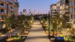 A well-lit pathway flanked by the modern NINE by Mirvac apartment buildings and greenery leads towards a distant city skyline at dusk, with illuminated skyscrapers against a twilight sky, marking the official opening of Henderson Park.