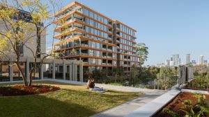 A person relaxes on a blanket in Henderson Park, nestled near the NINE by Mirvac apartment building with orange accents. The urban oasis offers views of the city skyline under a clear blue sky, and visitors are excited for its official opening.