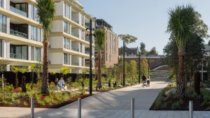 A modern residential complex, NINE by Mirvac, is adorned with palm trees and greenery lining a paved walkway. People sit on benches and stroll the path, savoring the sunny day. A set of stairs is visible in the background, hinting at Henderson Park's official opening nearby.