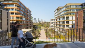 A man and woman stroll with a stroller and dog across a bridge overlooking NINE by Mirvac, a modern apartment complex. The scene features two multi-story buildings flanking a tree-lined pathway, with the city skyline in the background under the clear blue sky of Henderson Park.