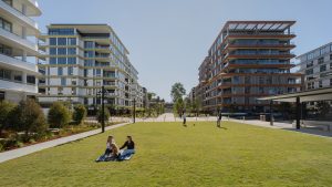 At Henderson Park, a spacious grassy area is embraced by the modern NINE by Mirvac apartments. Two people relax on a blanket with their dog in the foreground, while others stroll down tree-lined paths under a clear blue sky during its official opening ceremony.