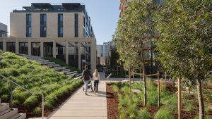 A modern urban landscape at Henderson Park features two people strolling along a pathway flanked by greenery and new architecture. The NINE by Mirvac complex, with its large windows and tiered seating, stands prominently under the clear blue sky during its official opening.