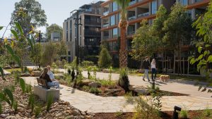 At the official opening of Henderson Park, people stroll with their dogs and relax on benches amidst modern apartment buildings, lush trees, and landscaped paths on this sunny day. The vibrant scene reflects the community spirit around NINE by Mirvac's newest urban oasis.