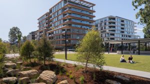 A modern apartment complex, part of the NINE by Mirvac development, stands with large glass windows against a clear blue sky. In front, at Henderson Park, two people sit and chat on the grassy area. Thoughtful landscaping with rocks and small trees complements a pathway weaving through the scene.