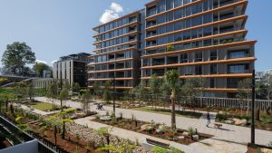 Modern apartment building with large windows and balconies, surrounded by landscaped gardens featuring walkways, trees, and small plants. A person strolls along the pathway on a sunny day at NINE by Mirvac during its official opening, under a clear blue sky near Henderson Park.