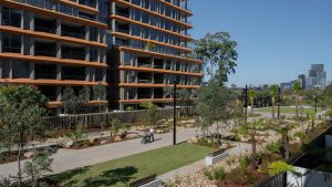 The NINE by Mirvac apartment building boasts modern design with expansive balconies, nestled beside a landscaped garden in Henderson Park. A person with a stroller strolls along a paved path, while city buildings loom under the clear blue sky during its official opening.