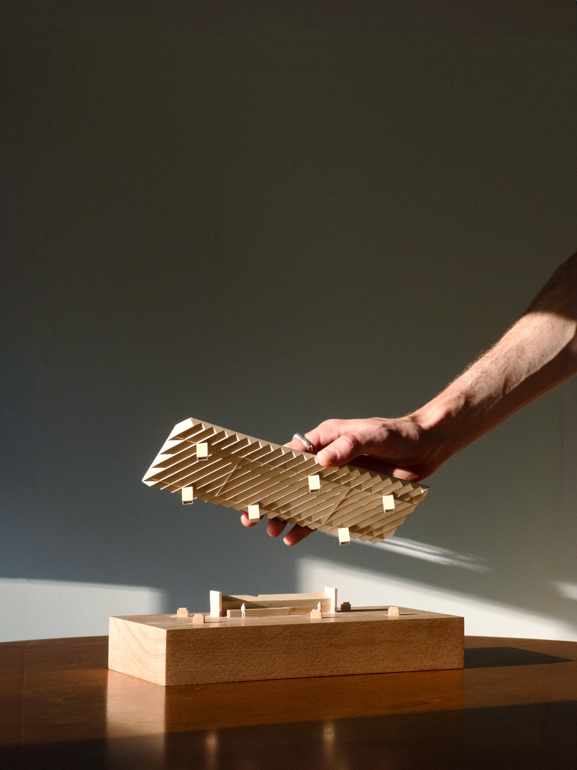 A person's hand holding a wooden architectural model, reminiscent of entries in a design competition, with layered rectangular pieces poised above a wooden base. The scene is brightly lit, casting shadows on a smooth, dark surface akin to the Sculpture Garden at the National Gallery of Australia.