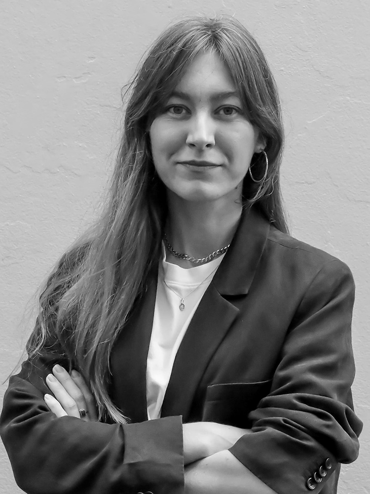 A woman with long hair stands with her arms crossed, exuding a confident expression in a blazer over a shirt. Large hoop earrings complement her poised demeanor against a plain, light-colored wall. This black-and-white image captures Holly Riddington’s elegant presence.