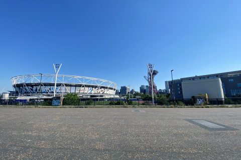 Wide-angle view of a sports stadium with a distinctive oval shape and white exterior at Queen Elizabeth Olympic Park. Nearby, a unique, tall red sculpture stands out against the clear blue sky. Buildings are visible in the background, with a large open paved area in the foreground.