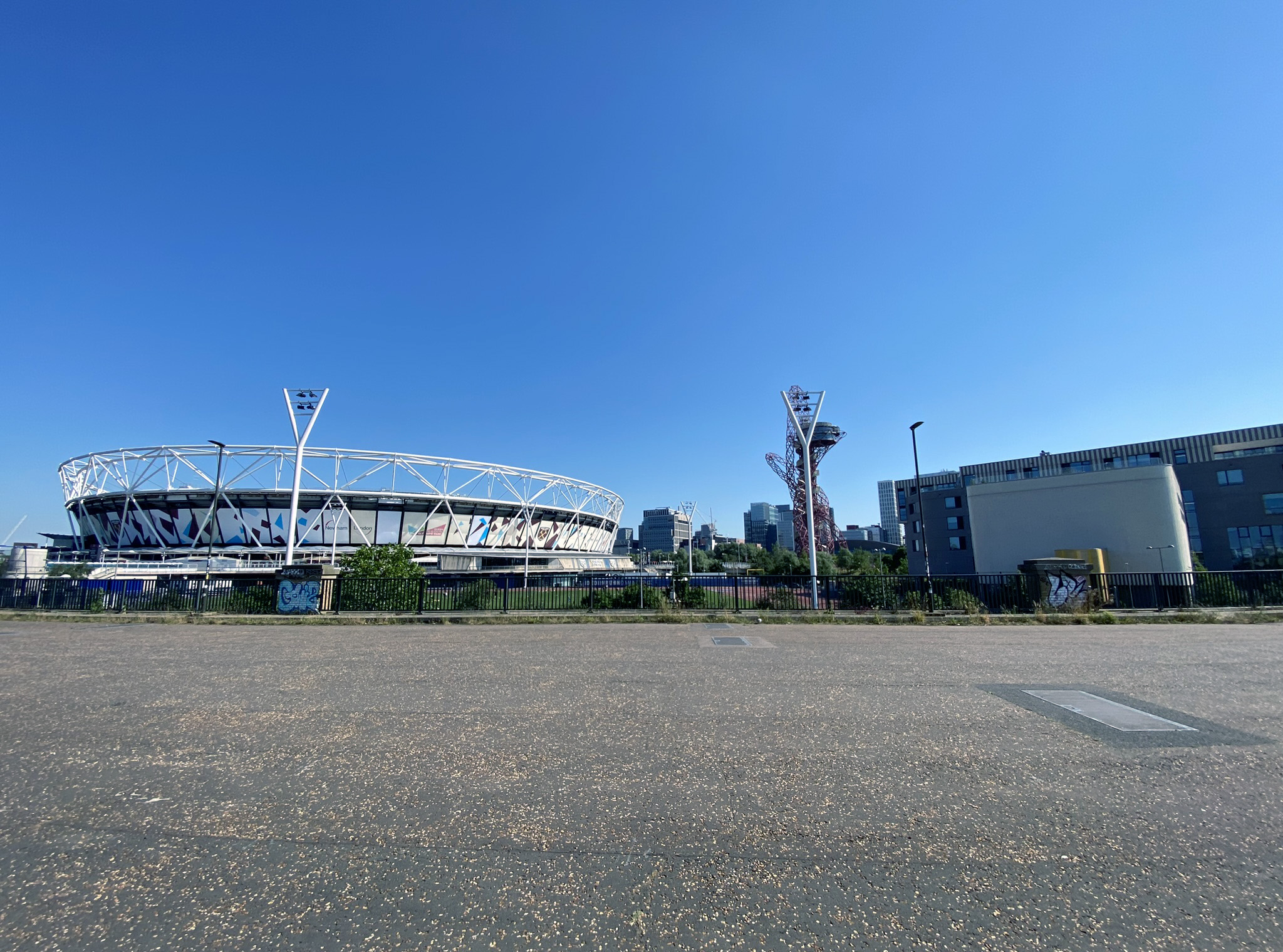 Wide-angle view of a sports stadium with a distinctive oval shape and white exterior at Queen Elizabeth Olympic Park. Nearby, a unique, tall red sculpture stands out against the clear blue sky. Buildings are visible in the background, with a large open paved area in the foreground.