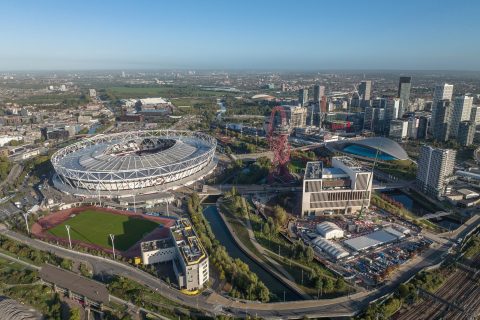 Aerial view of a large sports stadium, resembling the Olympic Park, surrounded by green fields and urban development. Nearby, a winding river, modern buildings by McGregor Coxall, and a unique red sculpture enhance the cityscape under a clear blue sky.