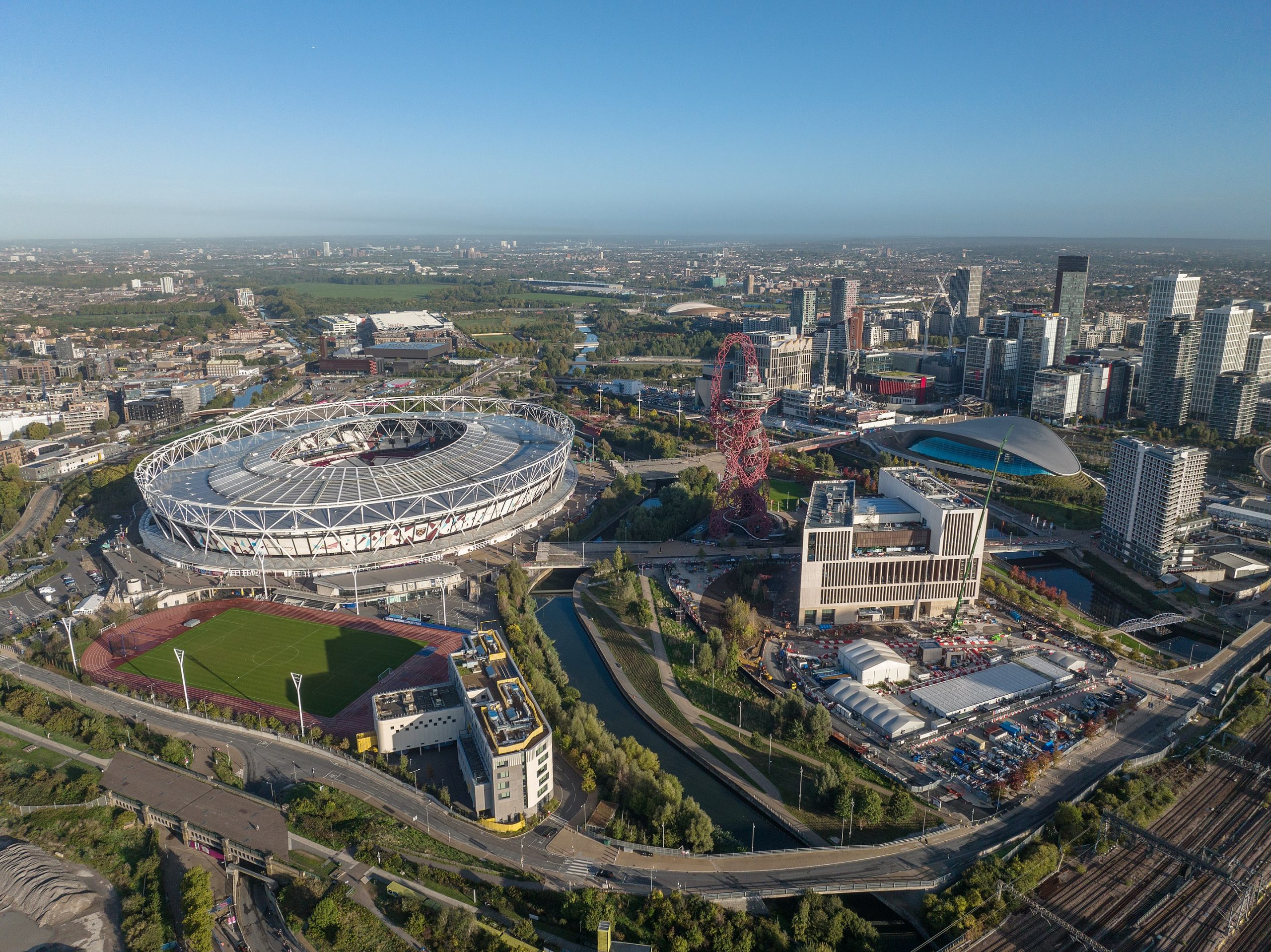 Aerial view of a large sports stadium, resembling the Olympic Park, surrounded by green fields and urban development. Nearby, a winding river, modern buildings by McGregor Coxall, and a unique red sculpture enhance the cityscape under a clear blue sky.