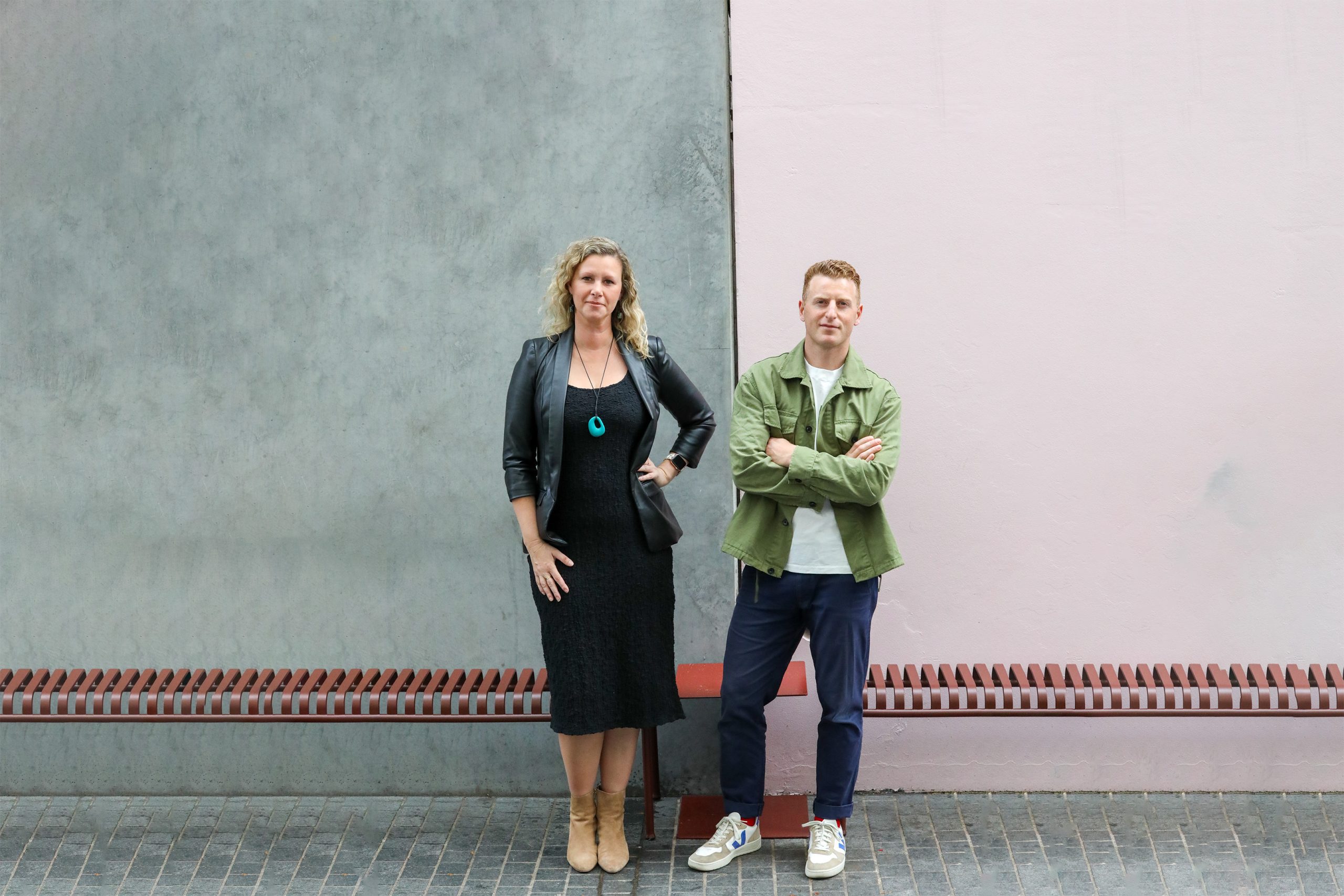 A woman and a man stand against a two-toned wall, with the left side gray and the right side light pink. The woman wears a black dress with a leather jacket, and the man wears a green jacket with jeans. Both look at the camera, their confident stance reflective of McGregor Coxall's new Managing Principals in the Sydney and Melbourne studios.