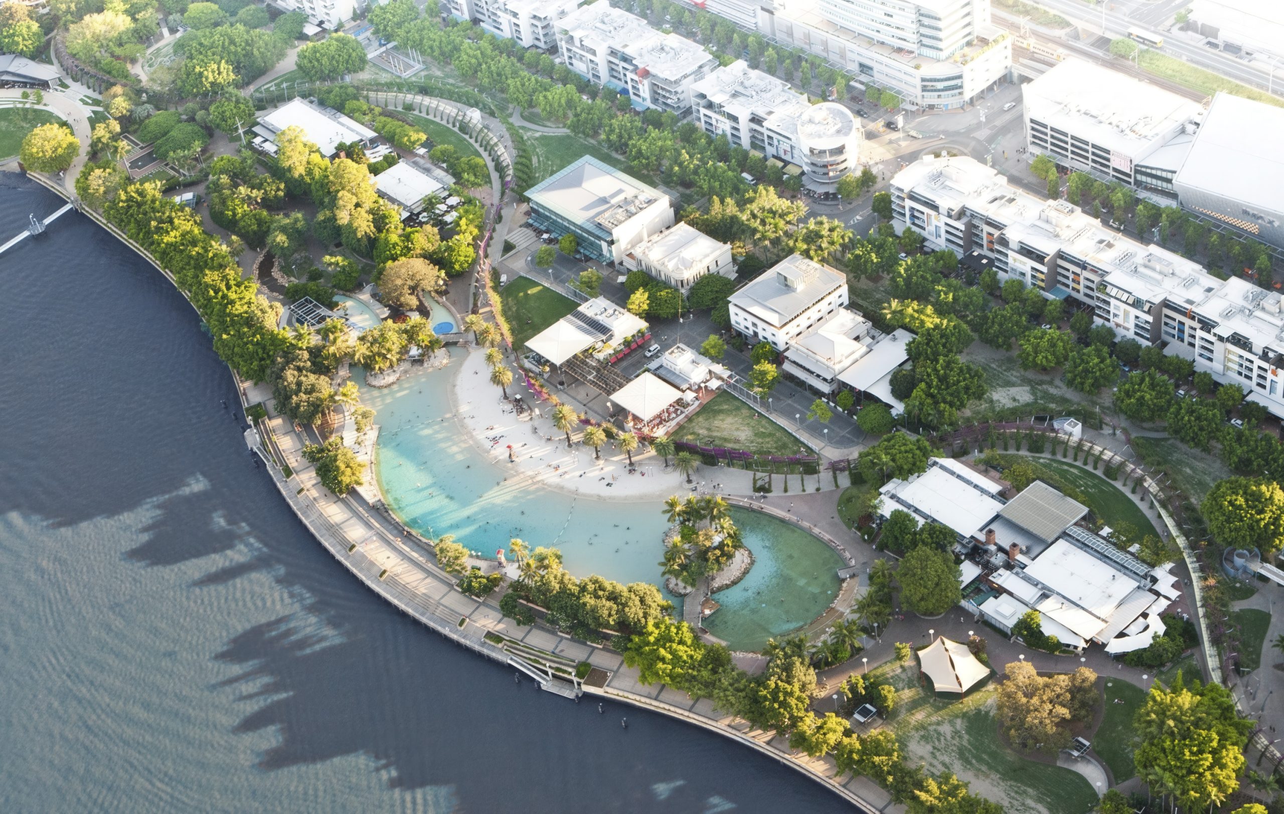 Aerial view of South Bank's riverside urban park featuring a lagoon-style pool nestled among lush trees and buildings. Walking paths weave through green spaces, inviting visitors to enjoy recreational activities. The park's design aligns with the area's biodiversity strategy, enhancing natural habitats.