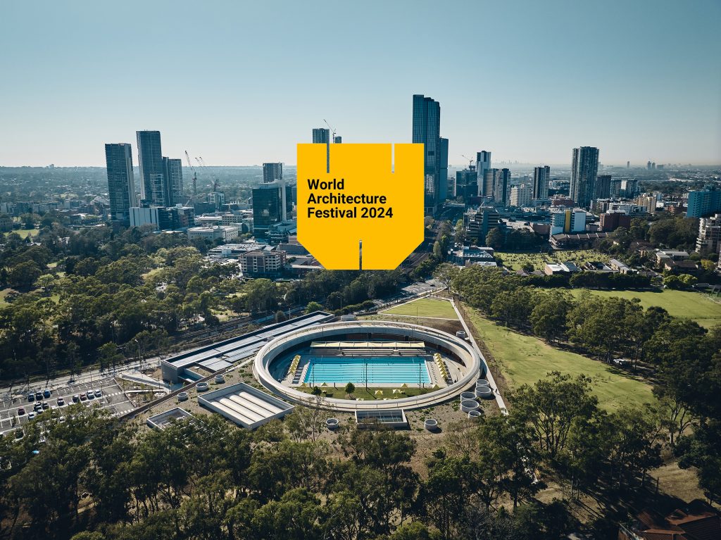 Aerial view of the circular Parramatta Aquatic Centre with a swimming pool, surrounded by trees and urban high-rises. A large yellow banner featuring 