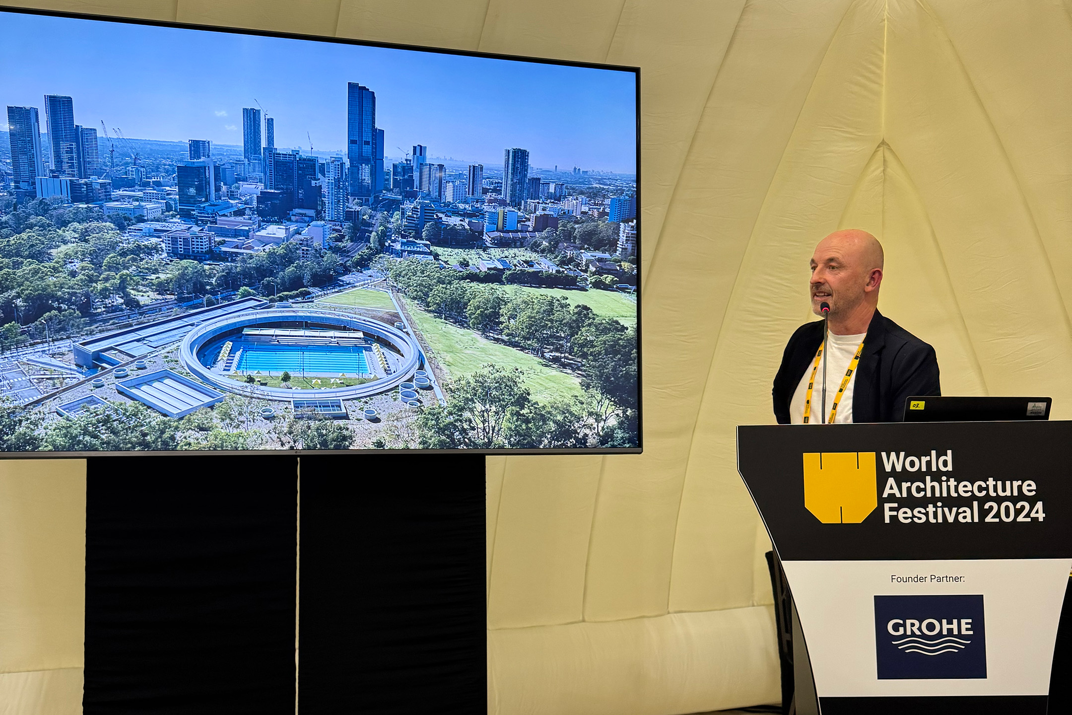 Andrew Burges stands at a podium labeled "World Architecture Festival 2024," presenting the innovative Parramatta Aquatic Centre. On a large screen, an aerial view of the stadium surrounded by trees and city buildings captivates the audience, hinting at its potential for the World Architecture Prize.