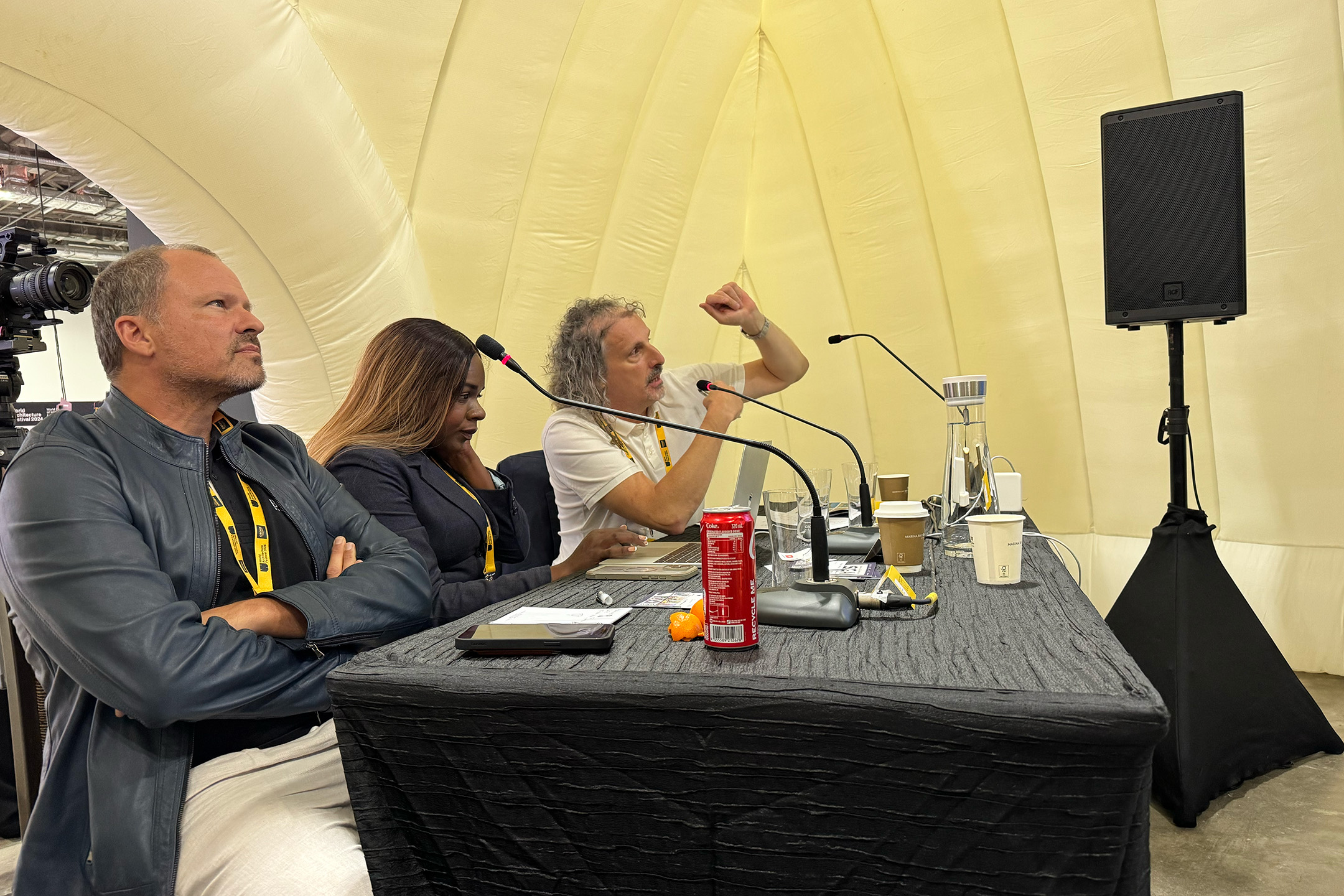 Inside the inflatable structure, three people sit at a table, microphones ready as they engage in a lively discussion about architecture. As one gestures passionately, cups and a red can scatter the table. The conversation flows seamlessly, reminiscent of sessions seen at the World Architecture Prize events.