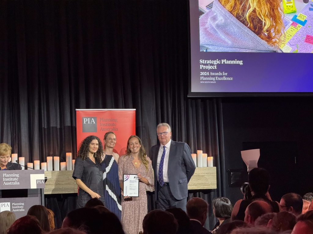 A group of four people stand on stage at an awards ceremony. Three hold a framed certificate, proudly acknowledging the PIA Commendation. Behind them is a banner with the Planning Institute Australia logo, while a presentation slide showcasing the Inner West Blue-Green Grid Strategy award winner is projected above.