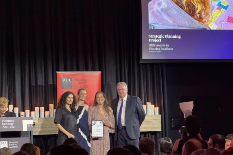 A group of four people stand on stage at an awards ceremony. Three hold a framed certificate, proudly acknowledging the PIA Commendation. Behind them is a banner with the Planning Institute Australia logo, while a presentation slide showcasing the Inner West Blue-Green Grid Strategy award winner is projected above.