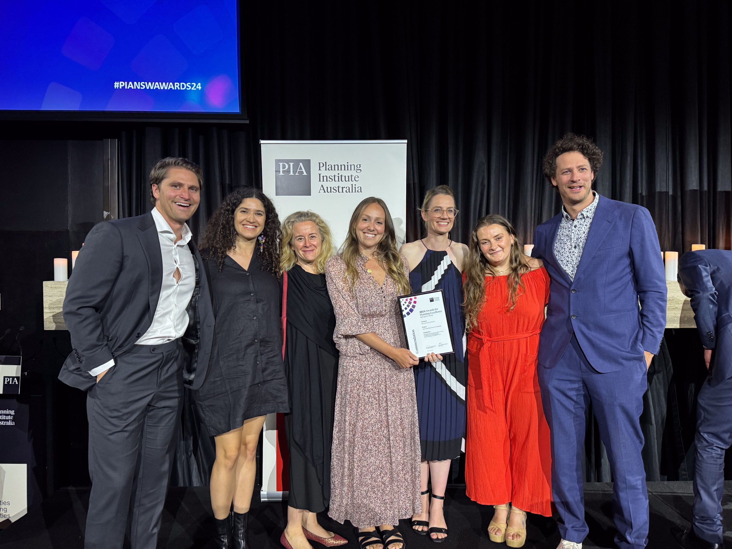 A group of seven people stand smiling together at an event, with one person proudly holding a PIA Commendation certificate. They are dressed formally, and there is a banner in the background for the Planning Institute of Australia.