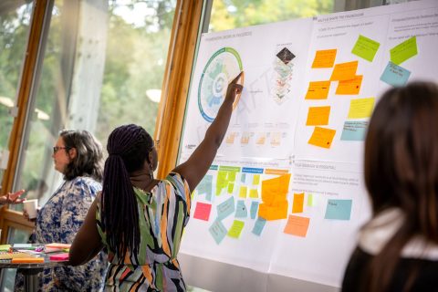 A woman with braided hair points at a wall covered in colorful sticky notes and charts during a brainstorming session on community flood resilience in a bright room. Another woman, an expert in groundwater, sits nearby, actively engaged in the activity.