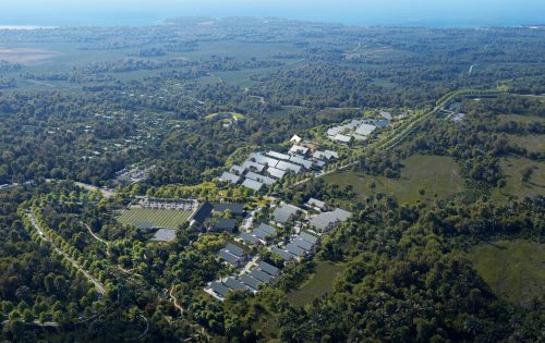 Aerial view of the Tropicalia Village masterplan reveals a large, modern complex with white-roofed buildings nestled among dense green trees, open fields, and a distant coastline under a clear sky.